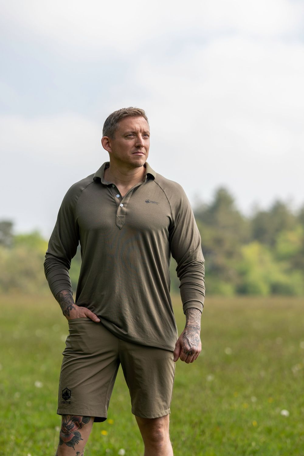 A person wearing an olive green polo and shorts stands in a grassy meadow on a sunny day.
