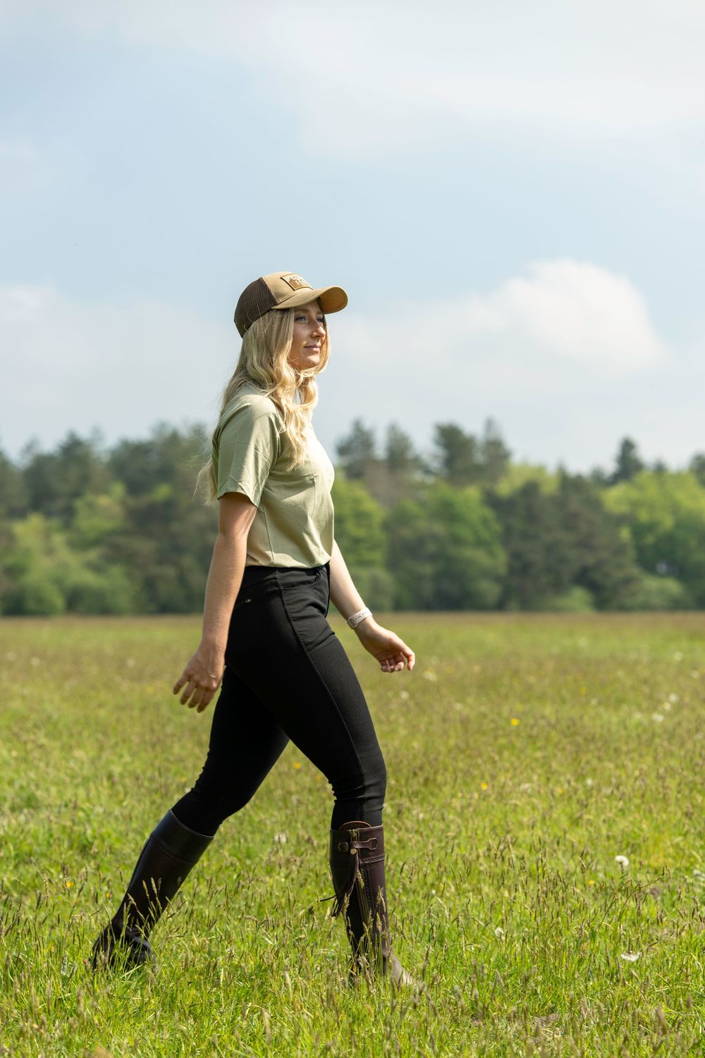 Someone in a tan baseball cap, light shirt and dark pants walks through a sunny meadow with trees in the background.