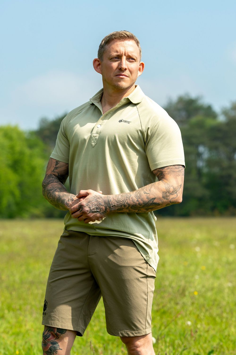 Athletic man in light polo shirt and khaki shorts standing in sunny meadow with tattoo-covered arms.