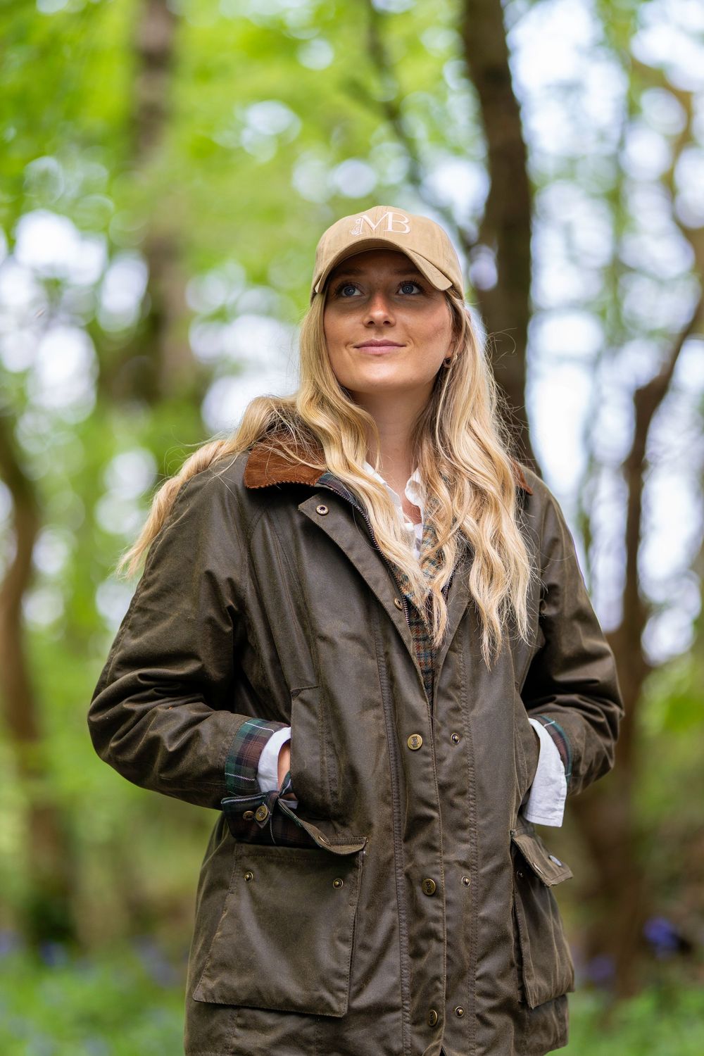 Person in a brown Barbour-style waxed jacket and baseball cap stands in a lush green forest setting.