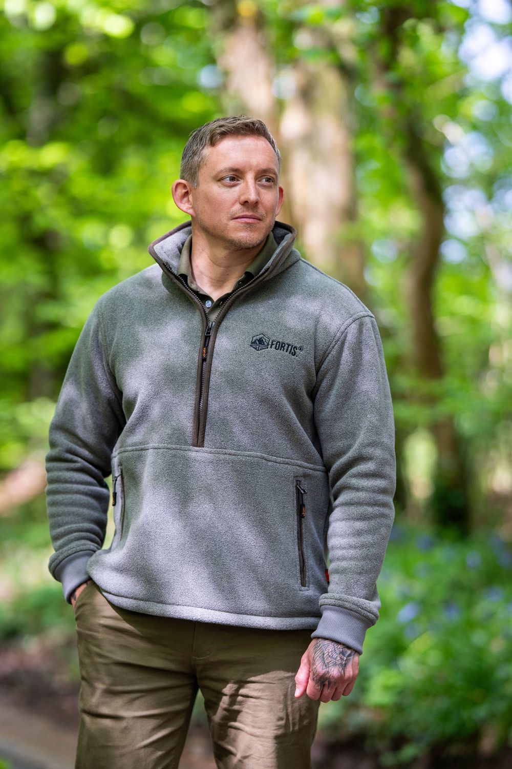 A person in a grey fleece pullover and brown pants stands outdoors in a lush green forest setting.