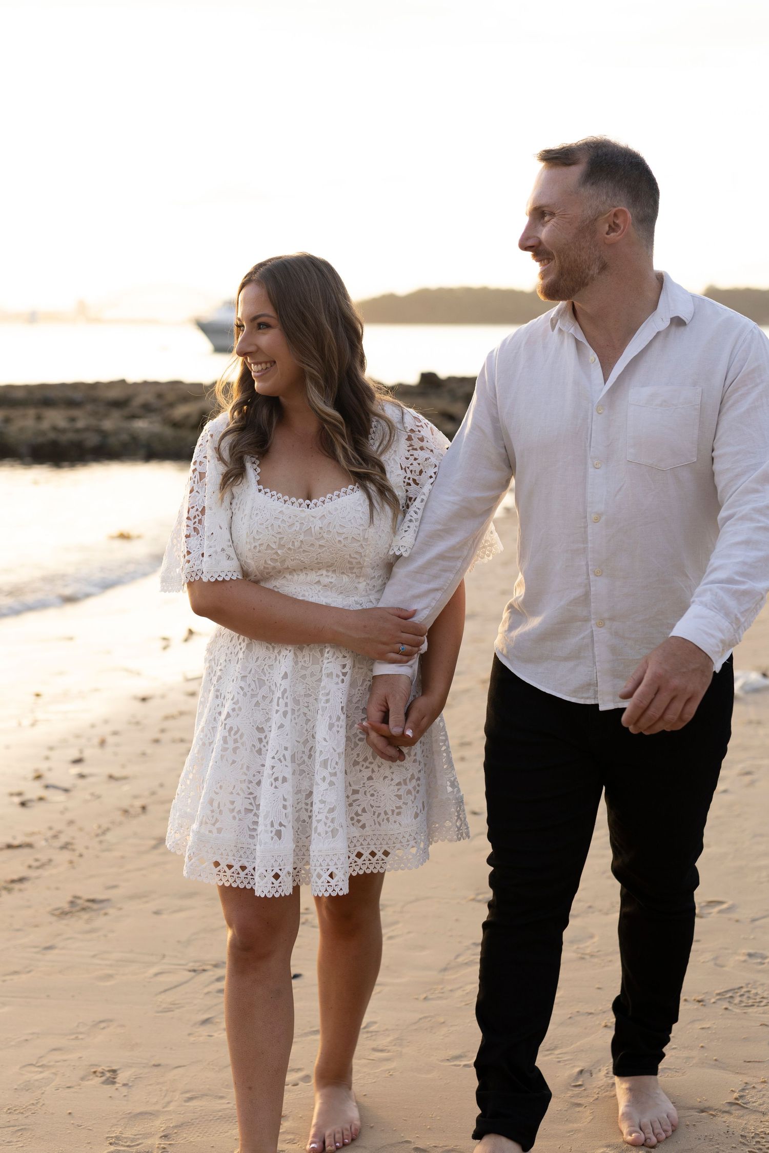 Romantic couple walking barefoot on beach at sunset with woman in white lace dress and man in white shirt.