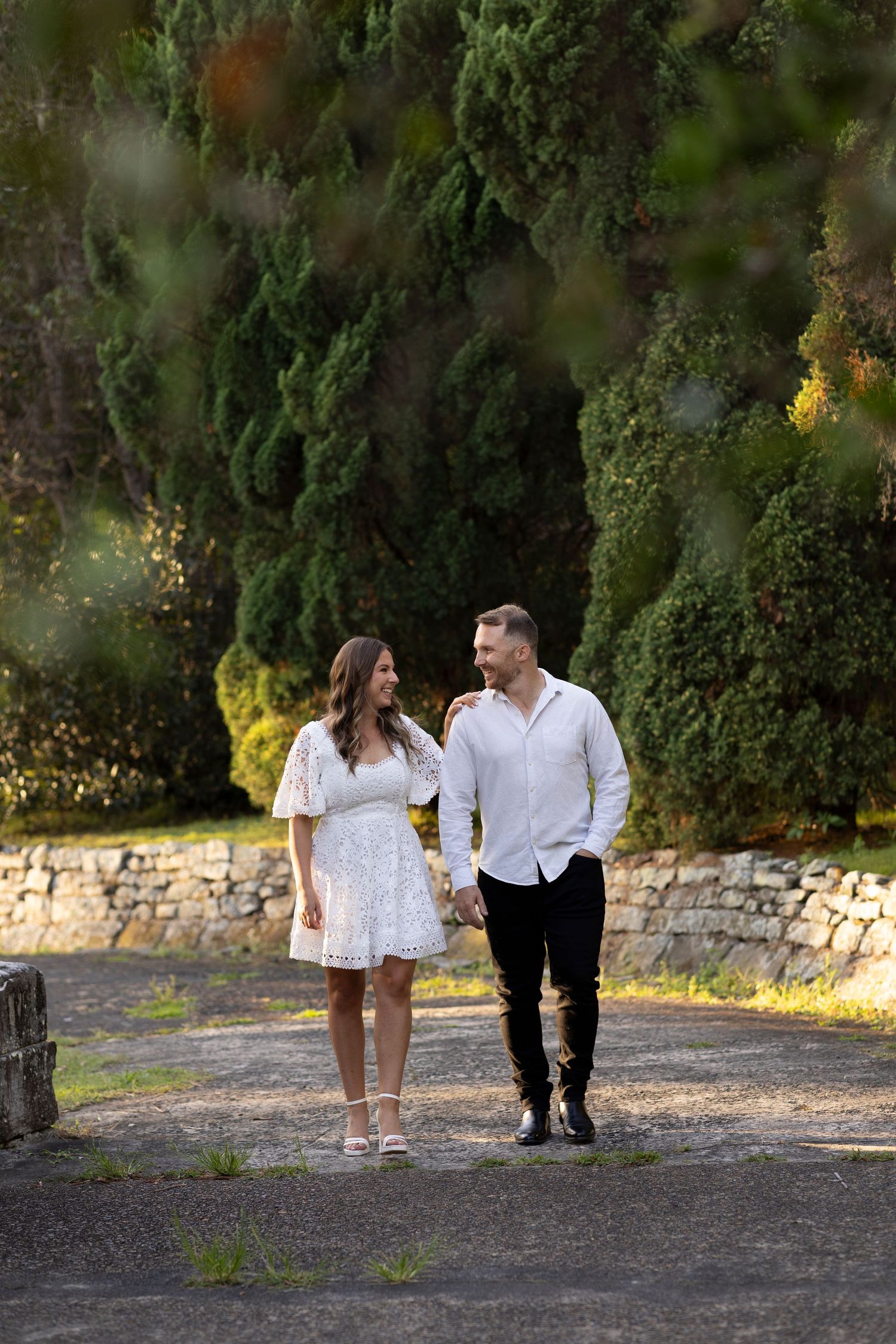 A couple walks together on a stone path surrounded by lush greenery and cypress trees on a sunny day.