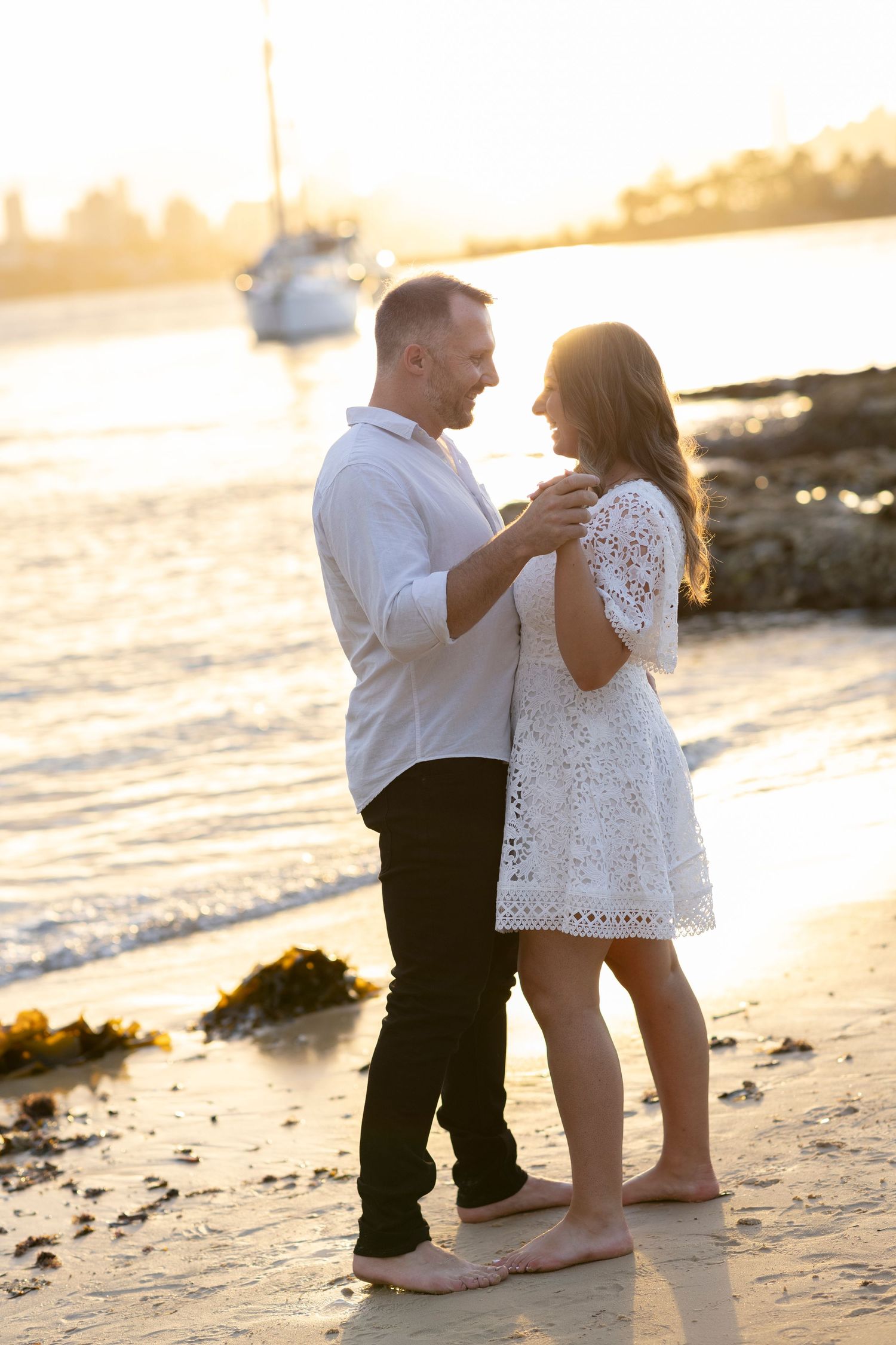 Romantic couple dancing on a beach at sunset with sailboats in background.