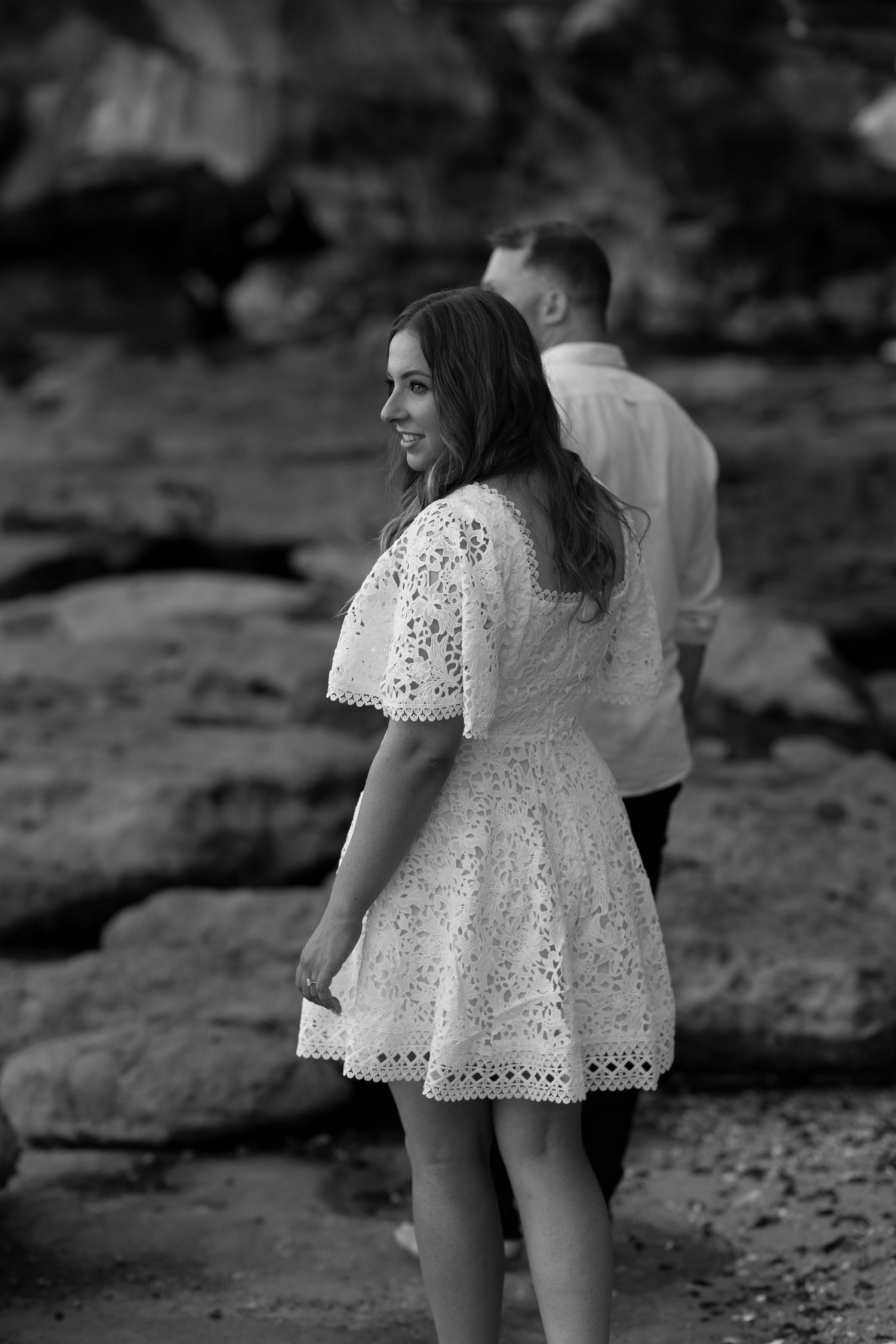 A couple on rocky terrain with the woman wearing a white lace dress looking back over her shoulder.