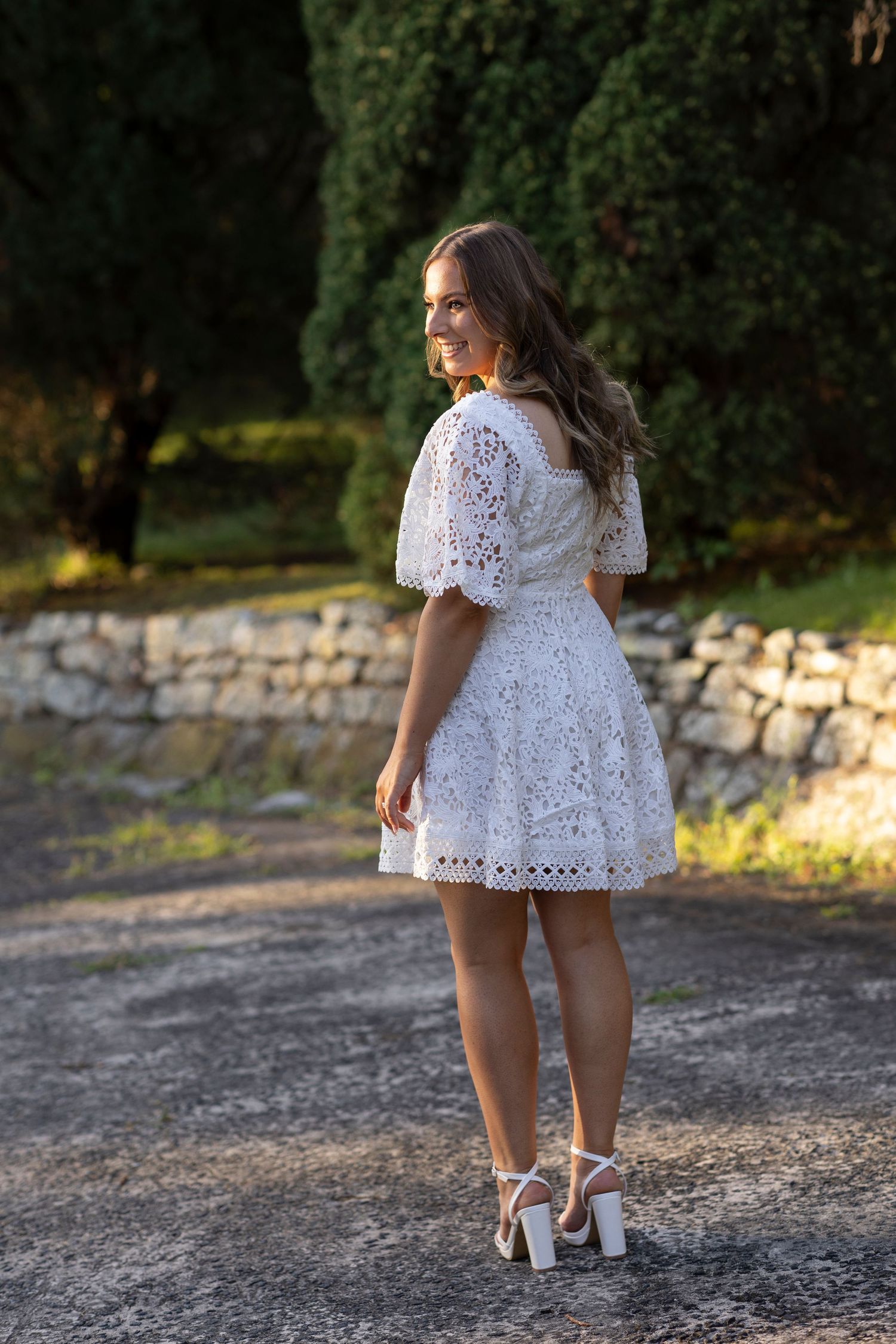Person in a white eyelet lace dress with puff sleeves and strappy heels poses outdoors near a stone wall.