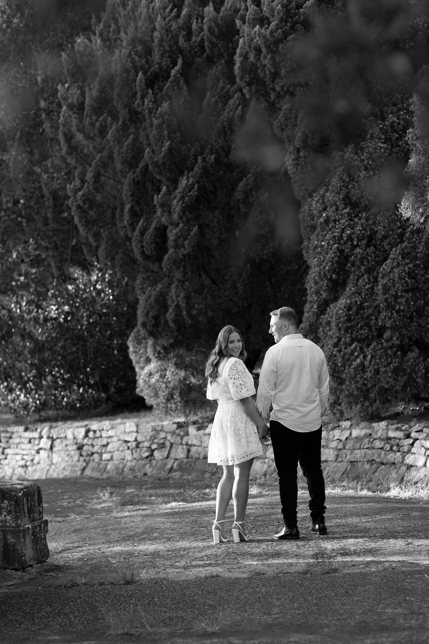 Two people walk together on a path beside a stone wall and trees in black and white.
