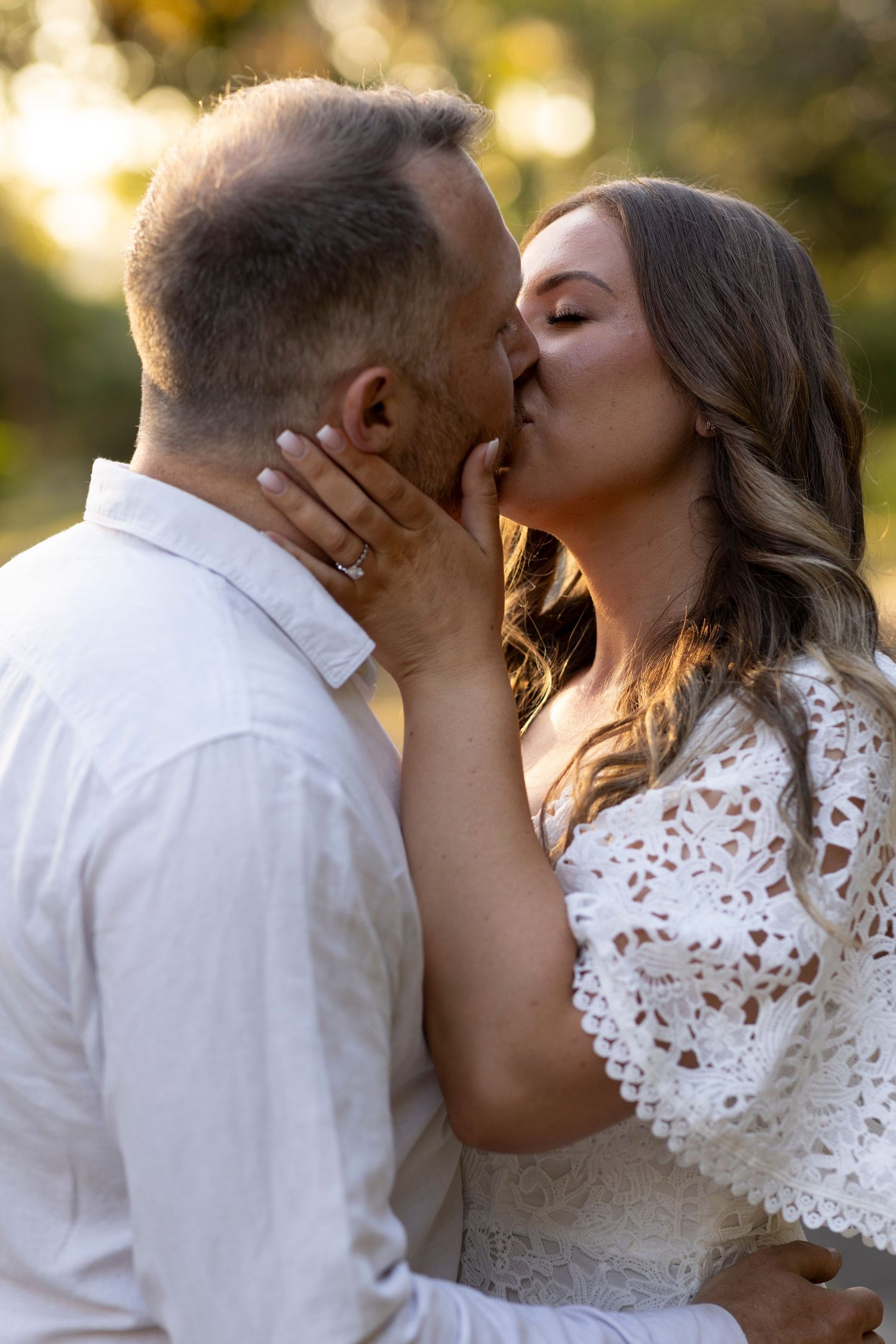 A romantic couple kiss outdoors in golden sunlight, the woman wearing white lace and man in light colored shirt.