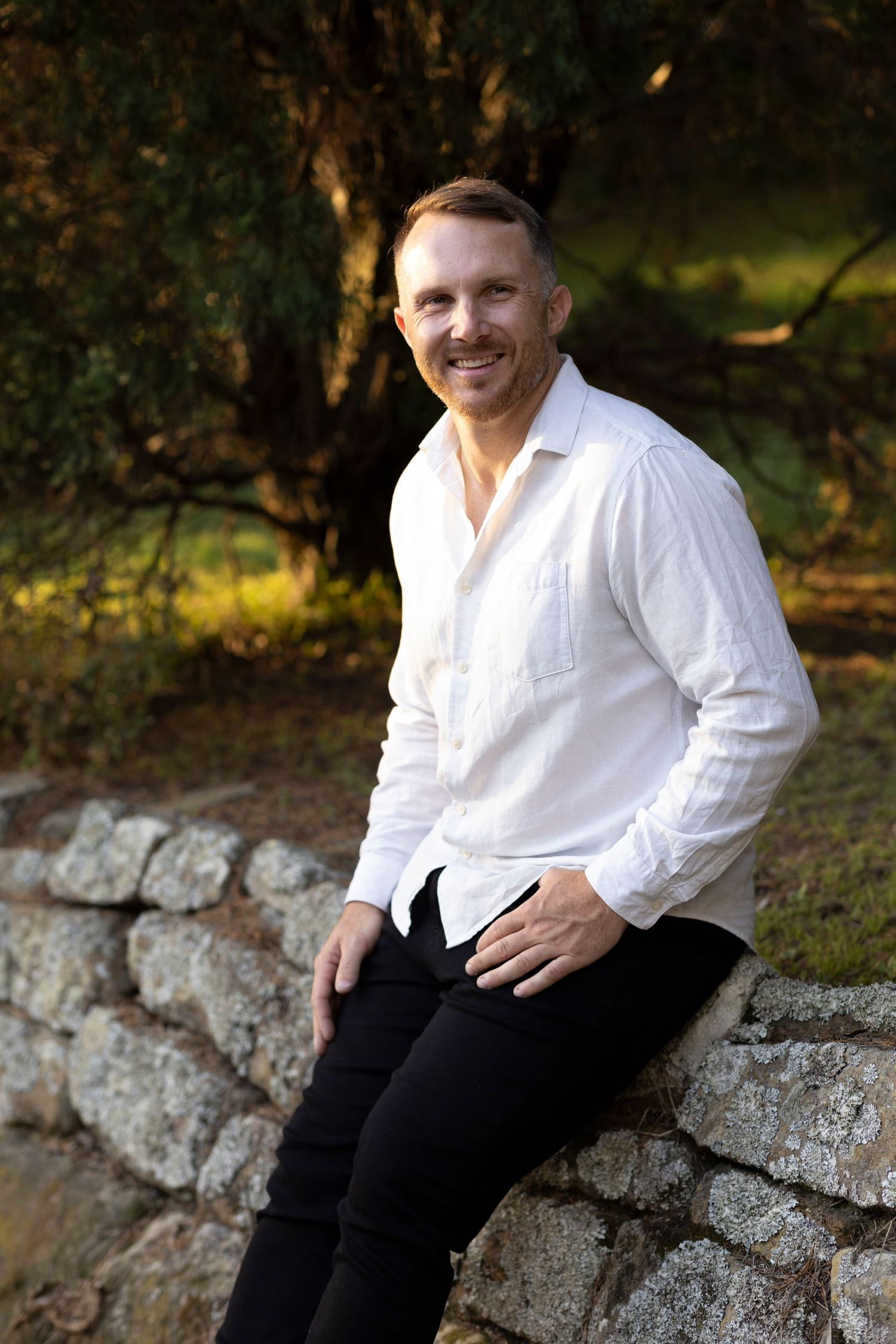 A person in a white button-down shirt and dark pants sits casually on a stone wall in a natural outdoor setting.
