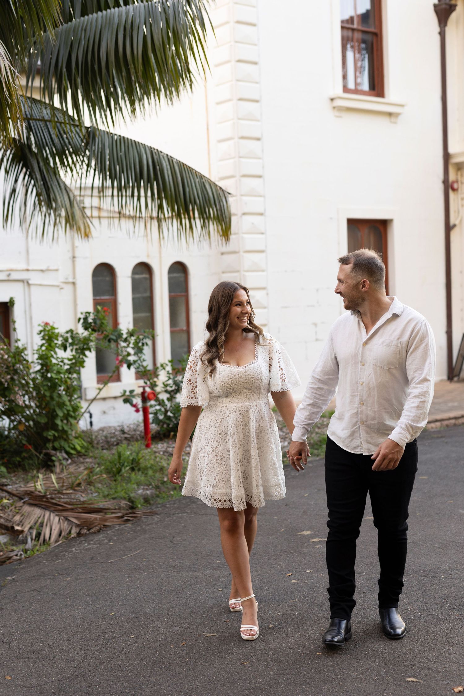 A couple walking together along a tropical pathway with palm trees and white building walls in the background.