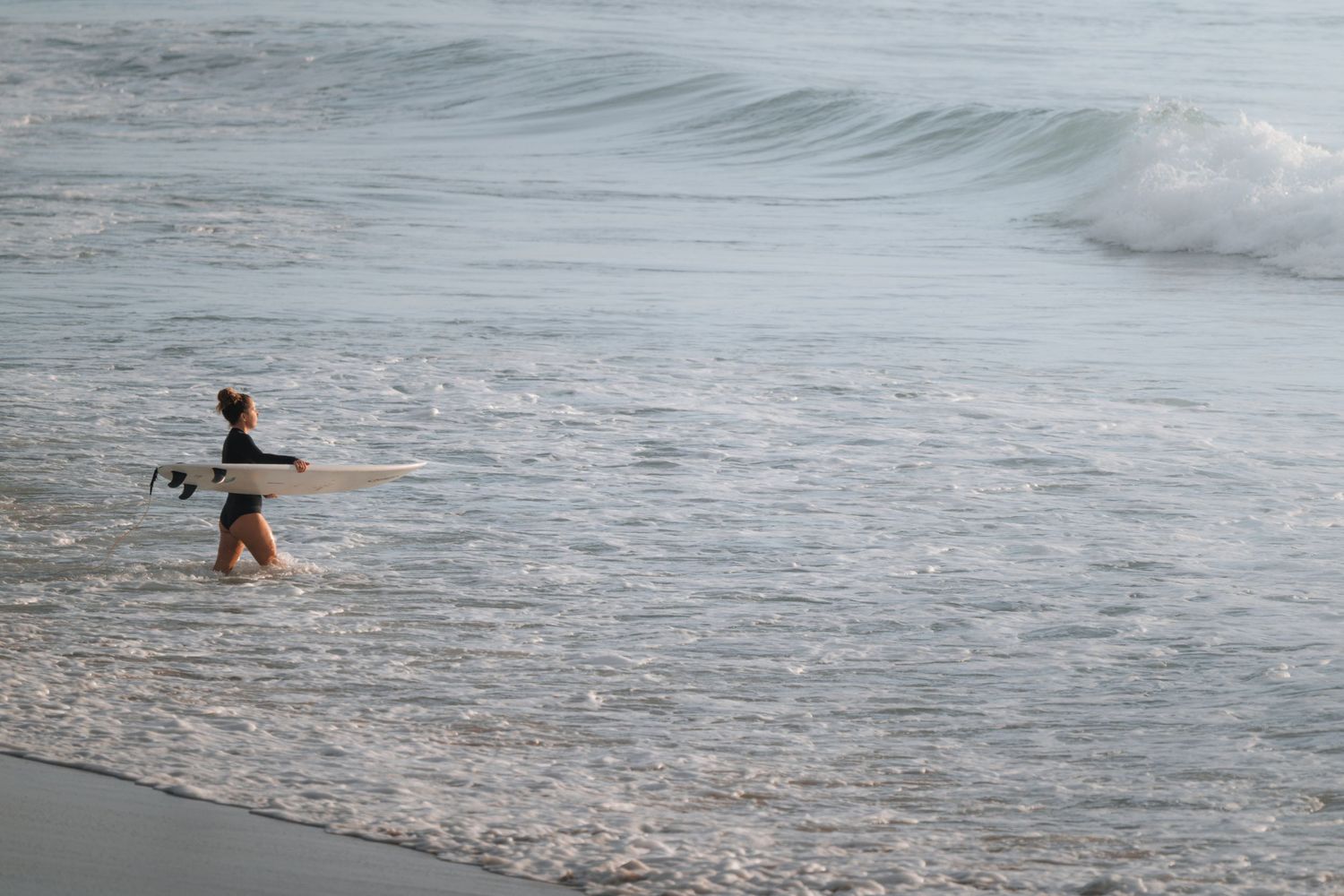 A surfer wades through the foamy waves along a misty beach shoreline.
