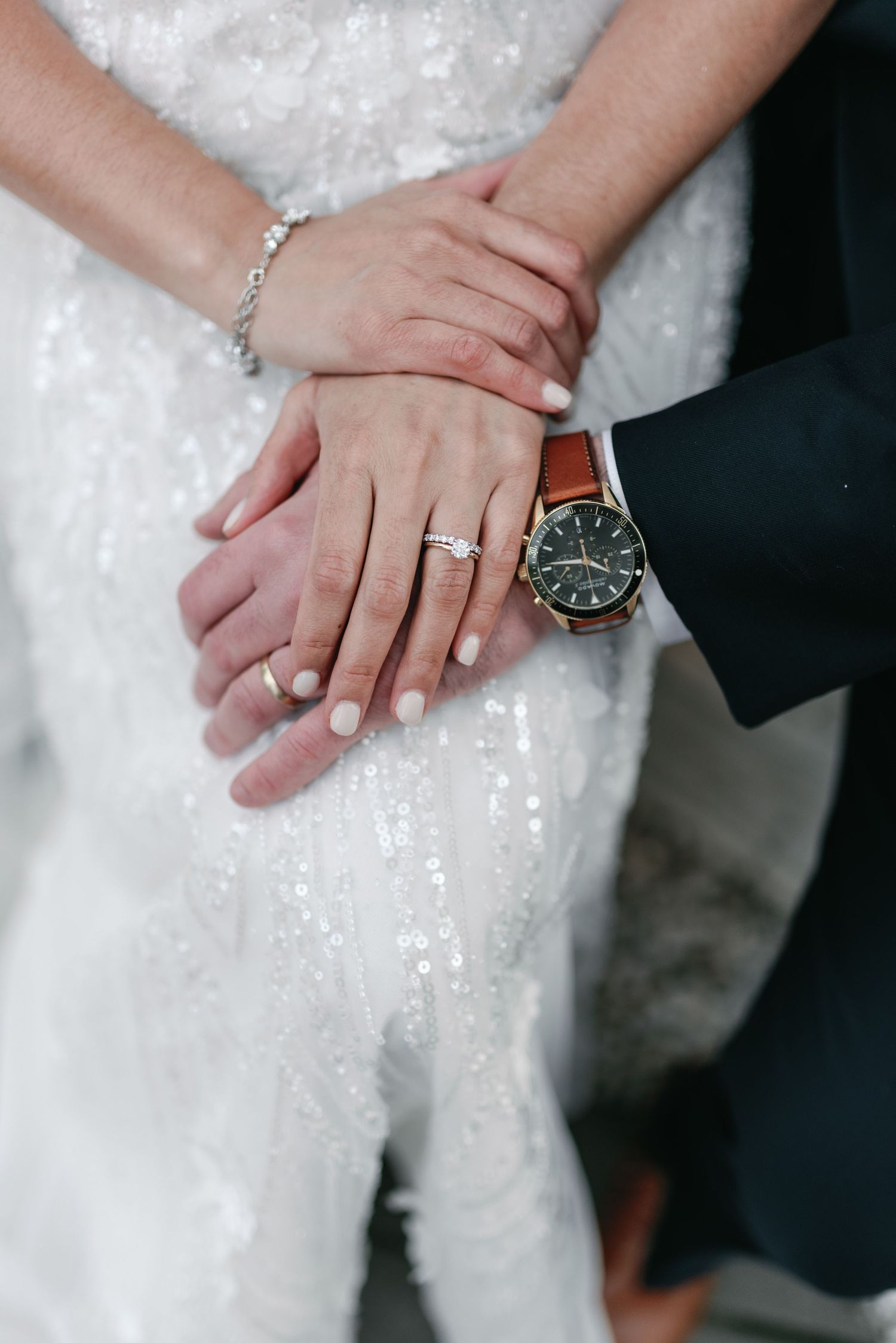 Close-up of newlywed couple's hands showing wedding ring, engagement ring, and groom's watch against white wedding dress.