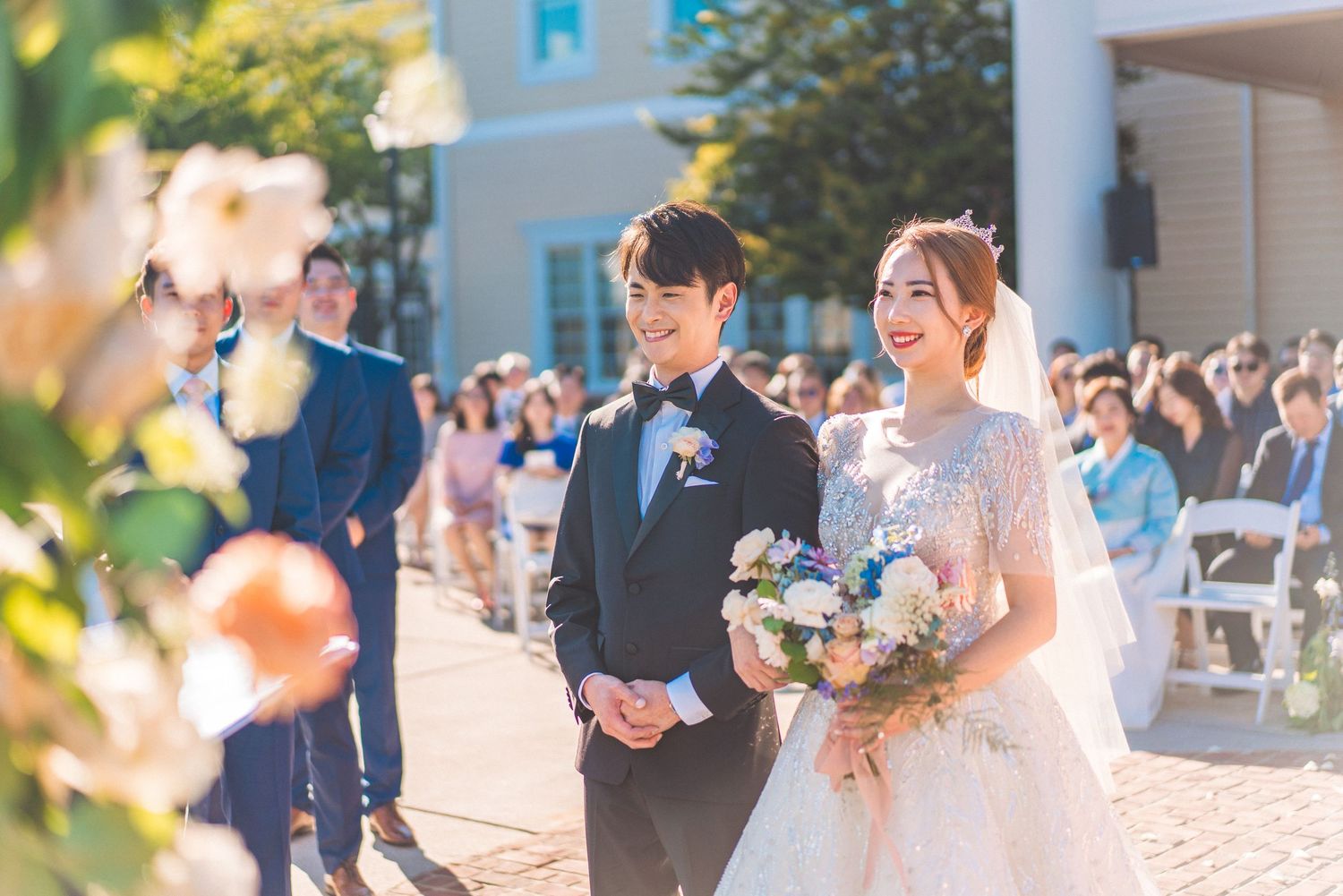 A wedding ceremony taking place outdoors with guests seated on white chairs.