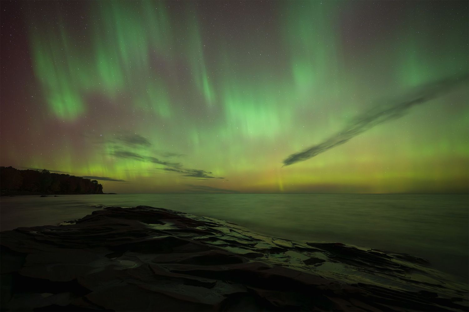 The Northern Lights dance across a dark sky above a rocky shoreline, casting vibrant green reflections on the water.