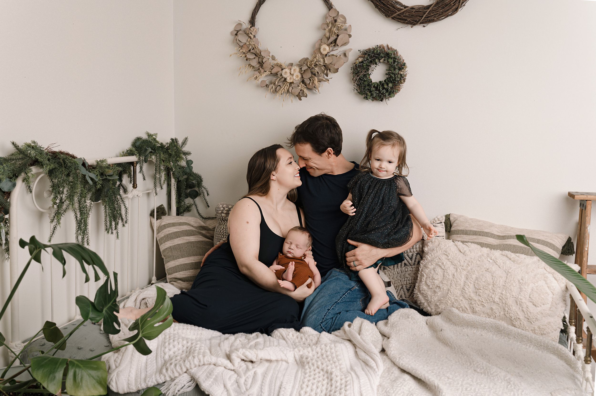 A family relaxes together on a cozy bed with white linens, surrounded by houseplants and decorative wall hangings.