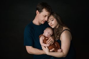 Portrait of tender family moment with parents cradling newborn baby against dark backdrop in studio setting.