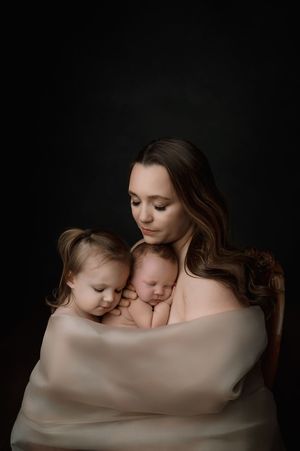 A tender studio portrait in dark tones shows two children cuddled against their mother, all wrapped in soft beige fabric.