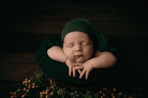 Sleeping newborn in emerald green wrap rests peacefully in round bowl surrounded by delicate woodland greenery.