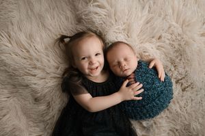 Two children lying on plush beige fur, one in black dress and one in teal outfit share sweet moment together.
