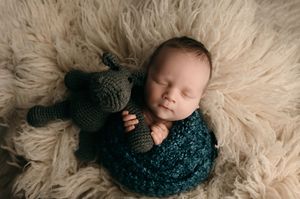 Sleeping newborn swaddled in teal knit wrap rests on fluffy beige blanket with dark plush teddy bear.