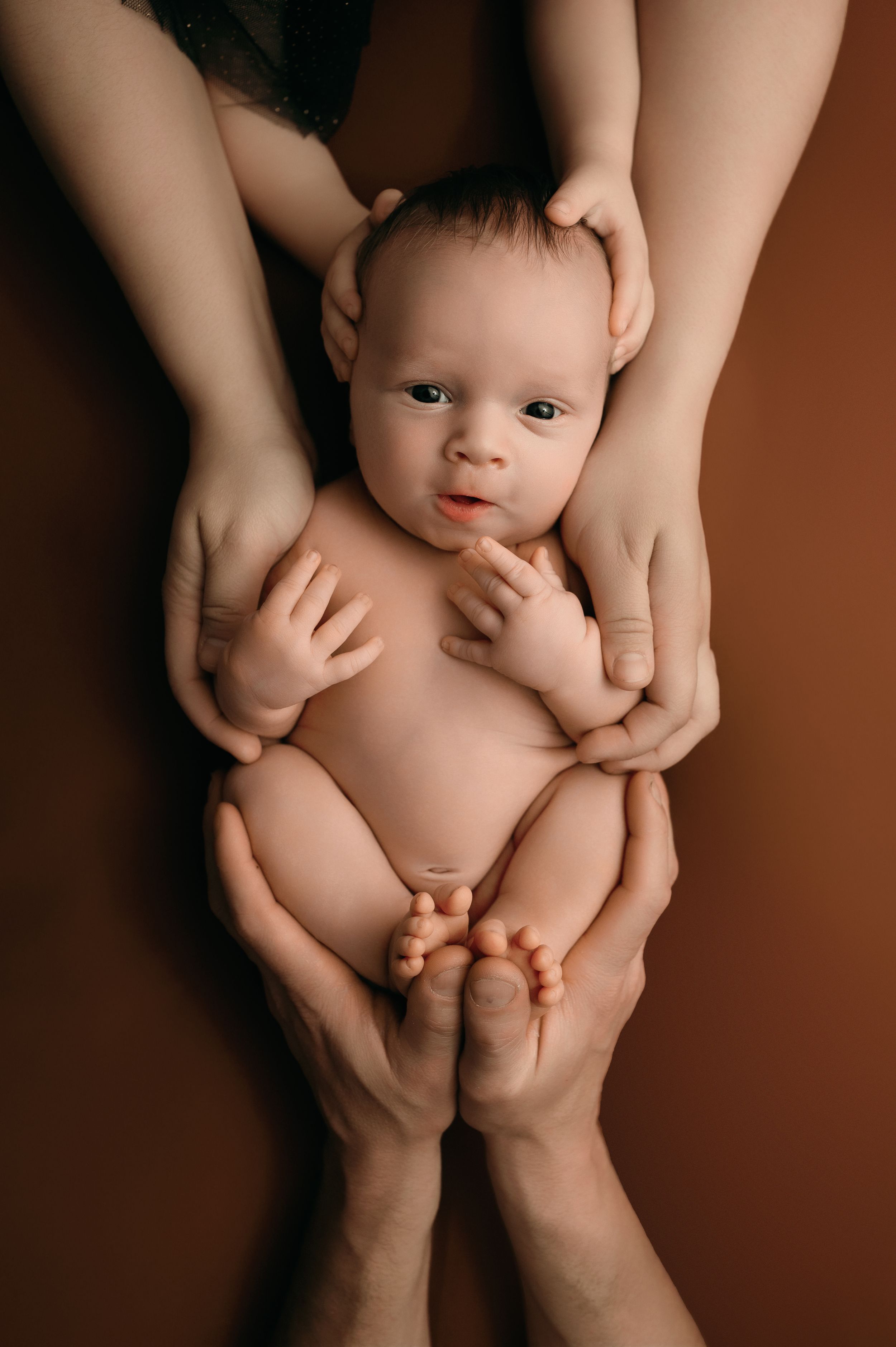 Caring hands gently hold a baby against a dark brown background in an intimate portrait photograph.