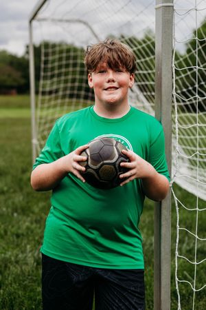 A young soccer player in a bright green t-shirt holds a black soccer ball while standing by a goal post on a grassy field.