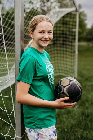 Young soccer player in green jersey holds black ball while standing by goal post on grassy field.
