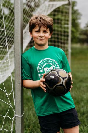 A young soccer player in a green jersey holds a black soccer ball while standing by a goal post.
