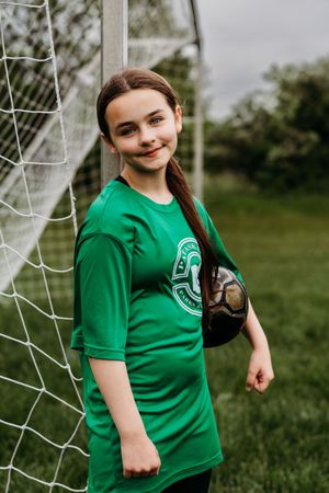 Soccer player in bright green jersey stands by goal net holding ball on grass field.