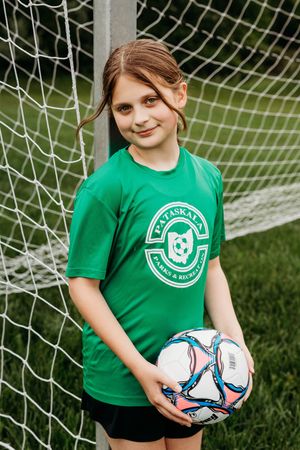 Young soccer player in a green team shirt stands by a goal net while holding a white and blue soccer ball.