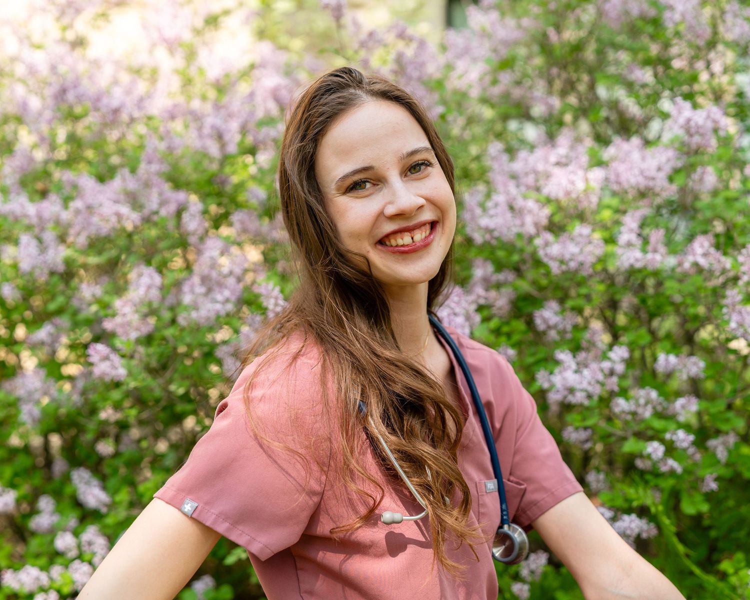 Healthcare professional in pink scrubs smiles warmly while standing among blooming spring flowers outdoors in nature.