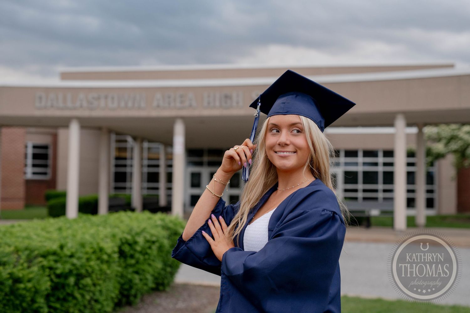 Graduate in cap and gown posing with diploma in front of school building on graduation day.