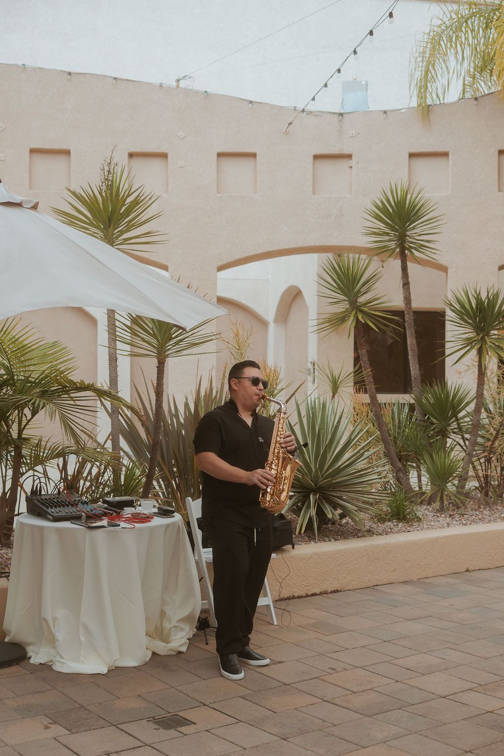 Saxophone musician performs at an outdoor venue with Mediterranean architecture and palm plants in the background.