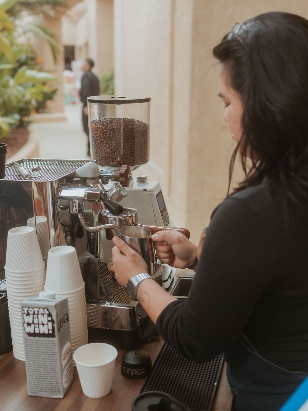 Barista operating an espresso machine with coffee beans in dispenser and disposable cups on counter.