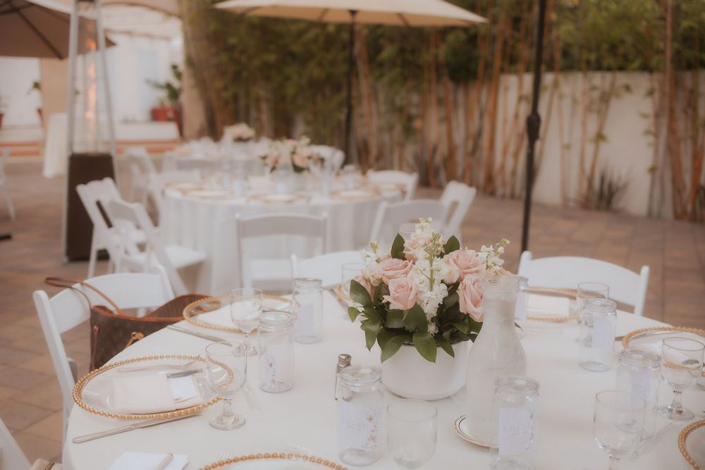 Elegant outdoor baby shower reception setup with white tables, chairs and delicate pink floral centerpieces under a patio umbrella.
