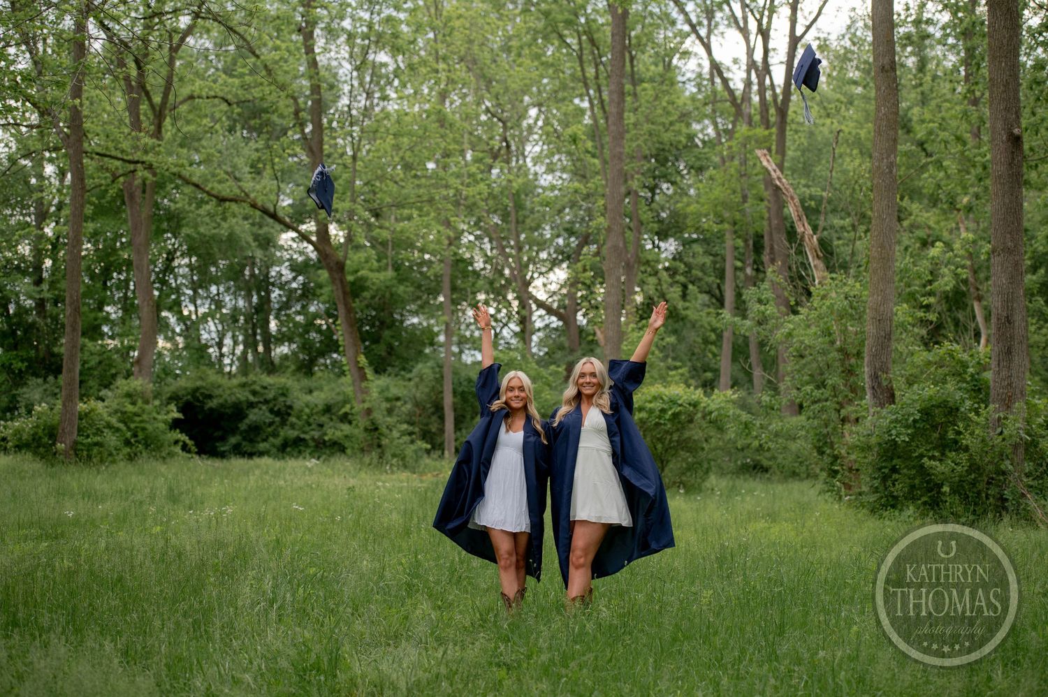 Two figures in navy blue graduation gowns celebrate with arms raised in a lush green meadow surrounded by trees.