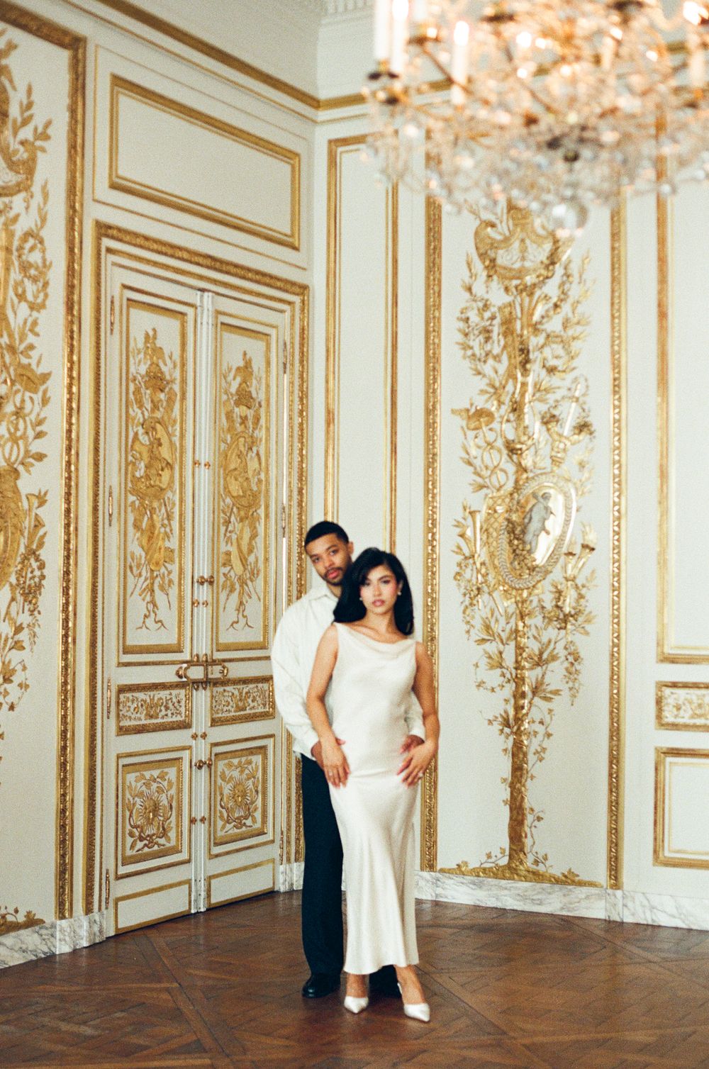 A romantic couple poses in an ornate room with gilded baroque wall panels and a crystal chandelier.