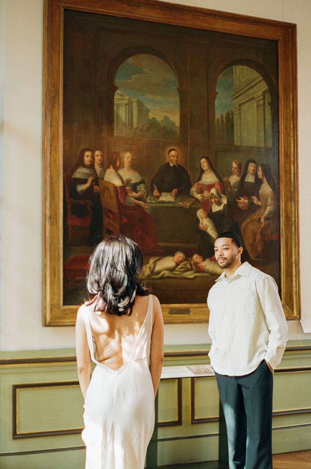 A couple admires a classical painting in an ornate room with decorative wood paneling.