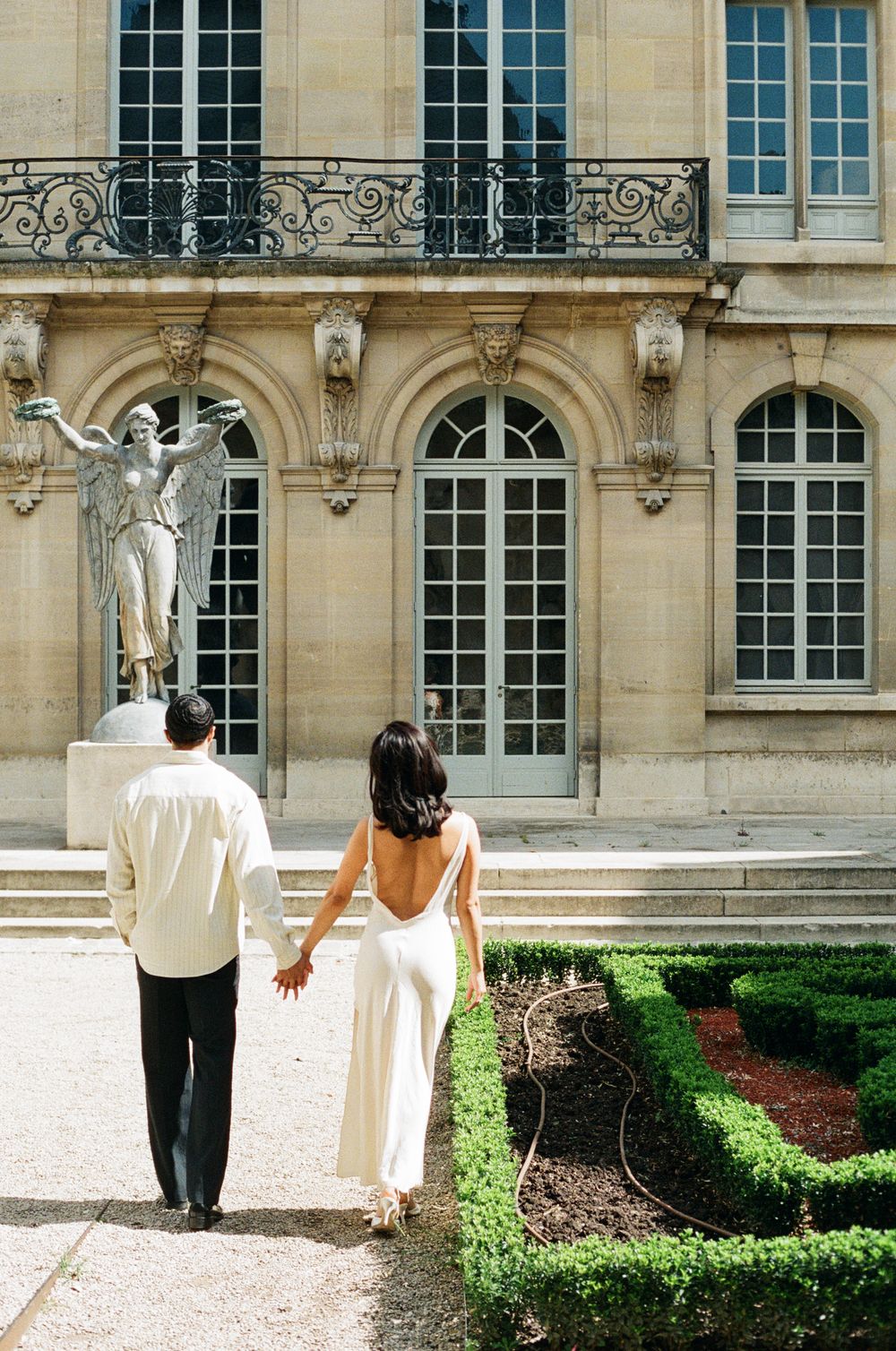 A couple in white clothing walks hand in hand through a formal garden with classical architecture.