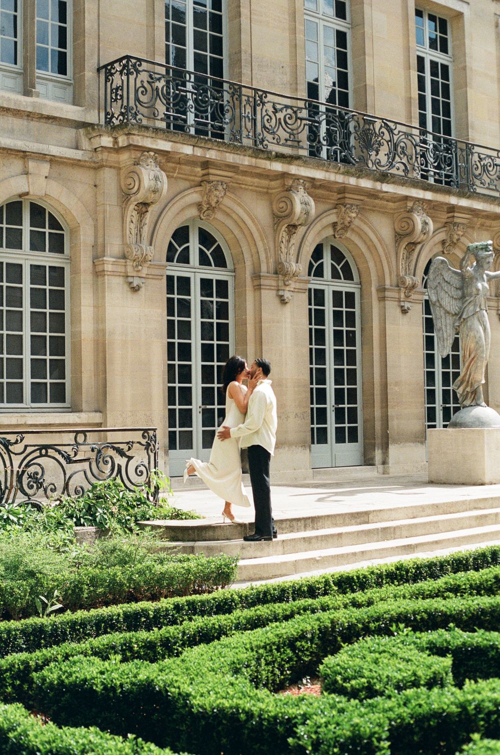 A couple embraces on the steps of a grand French-style mansion with ornate balconies and manicured gardens.