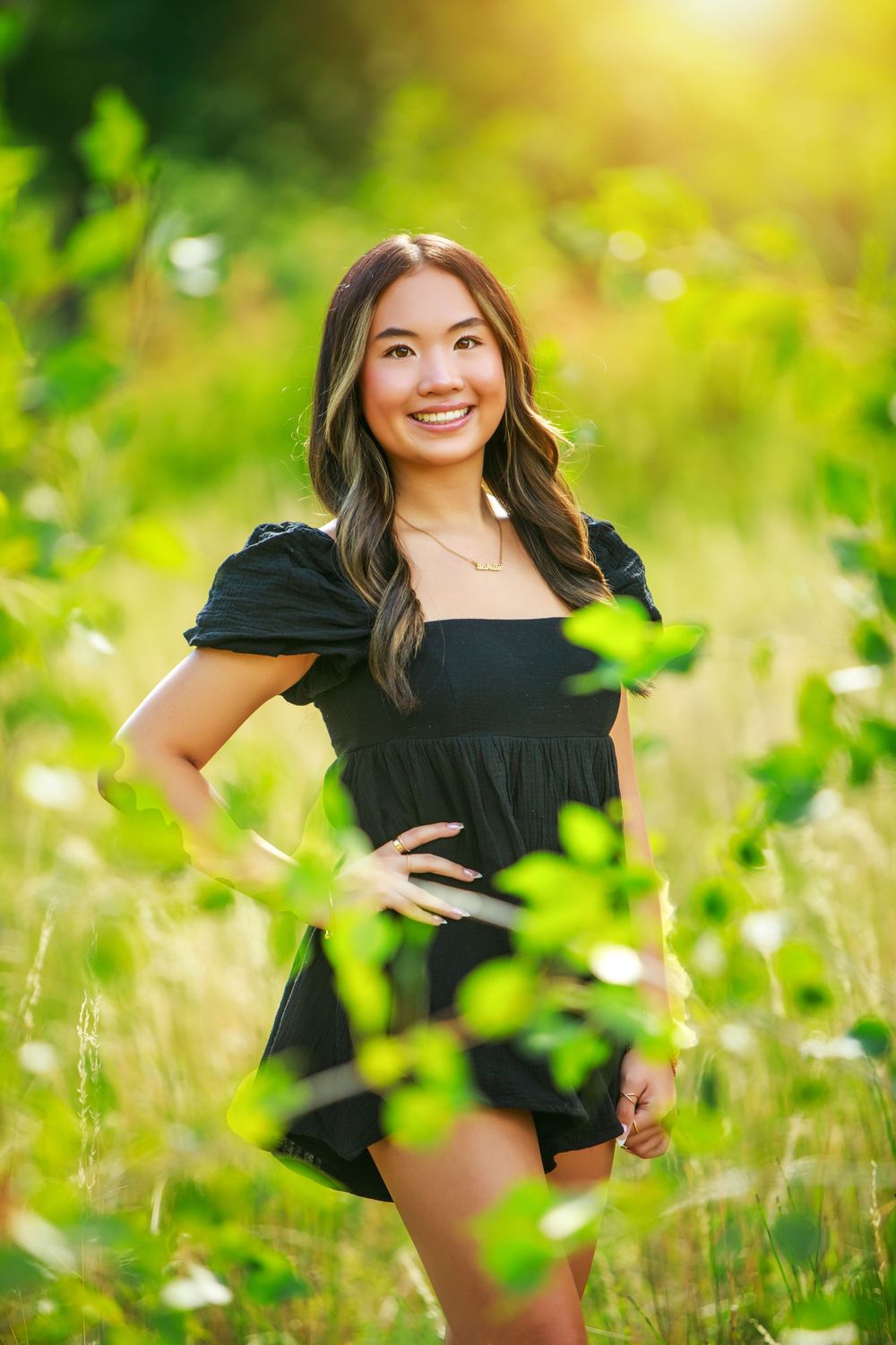 A joyful woman in a black dress poses in a sunlit meadow with green foliage and bokeh effects.