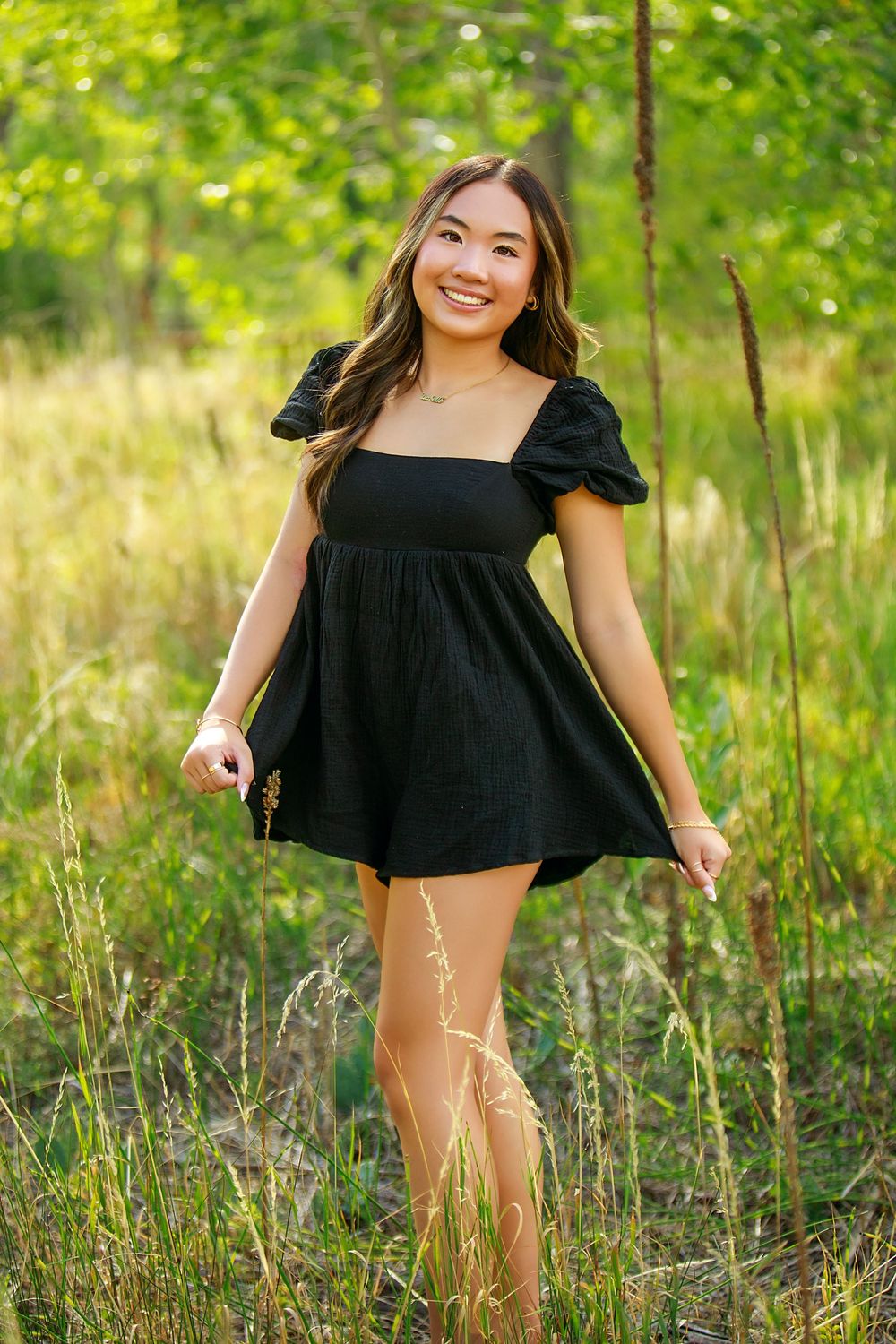 Person in a black summer dress poses playfully among tall grass and trees in a sunlit outdoor setting.