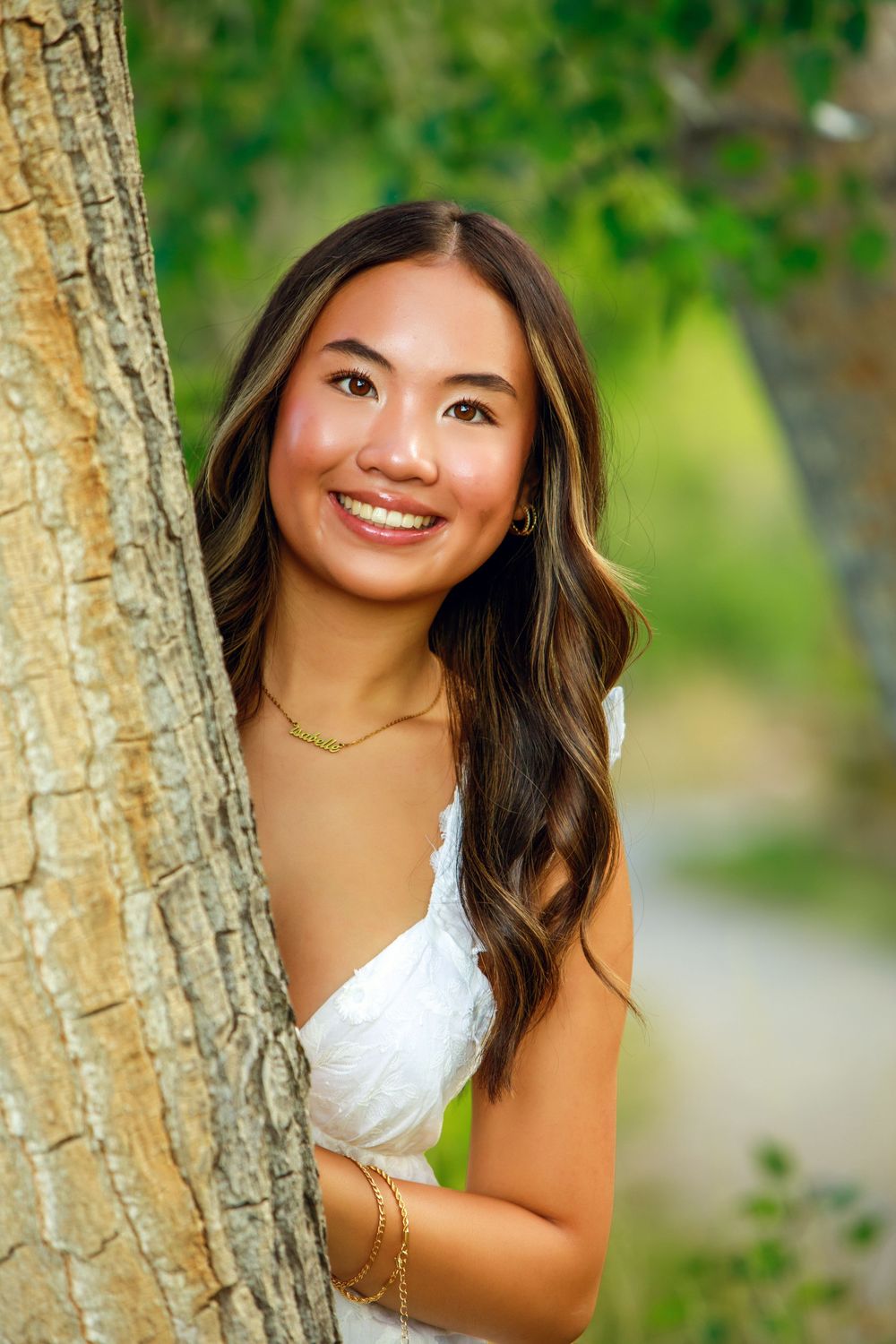 Portrait in a white dress next to a tree trunk with lush green foliage in the background.