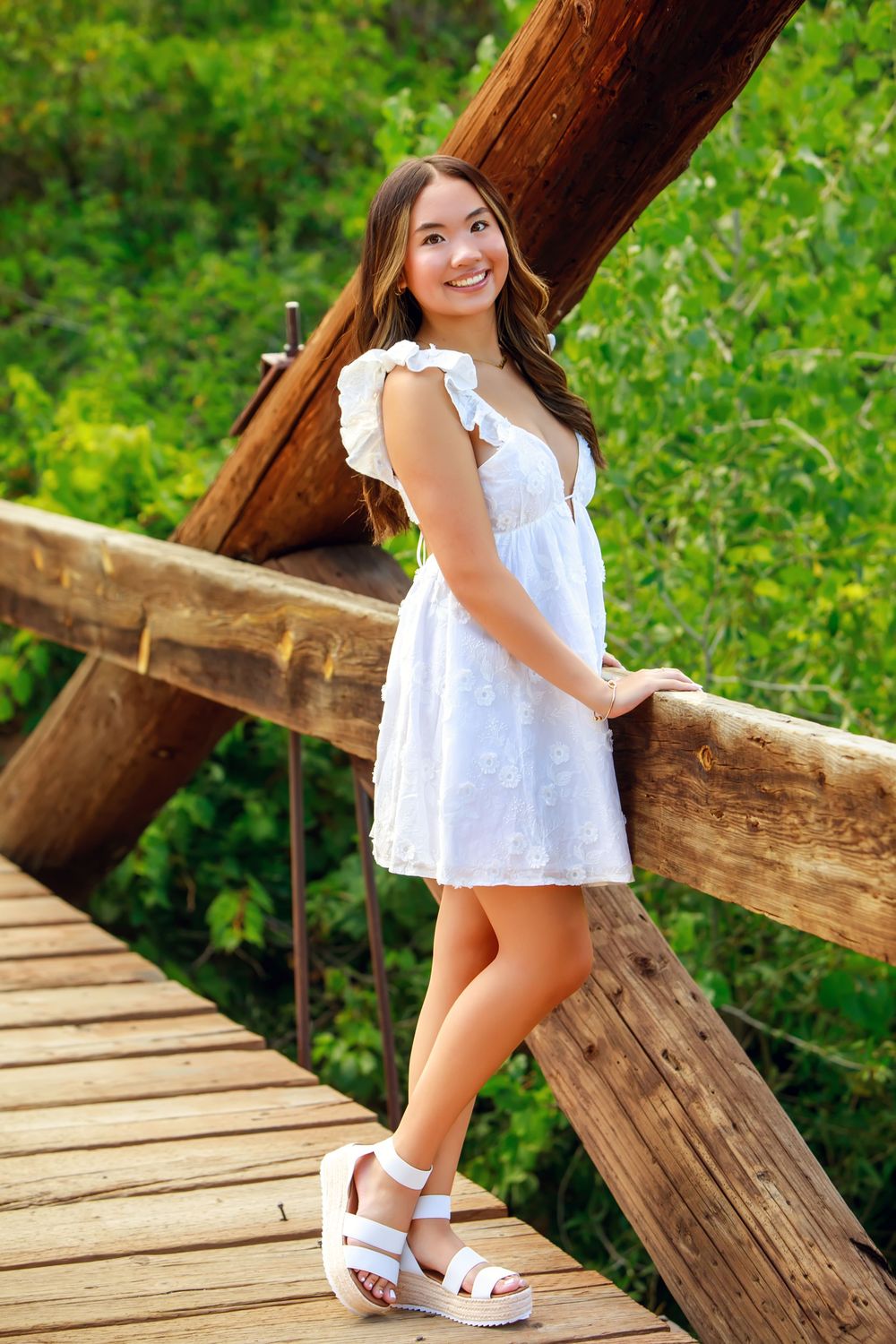 A smiling woman in a white sundress stands on a wooden bridge surrounded by lush greenery.