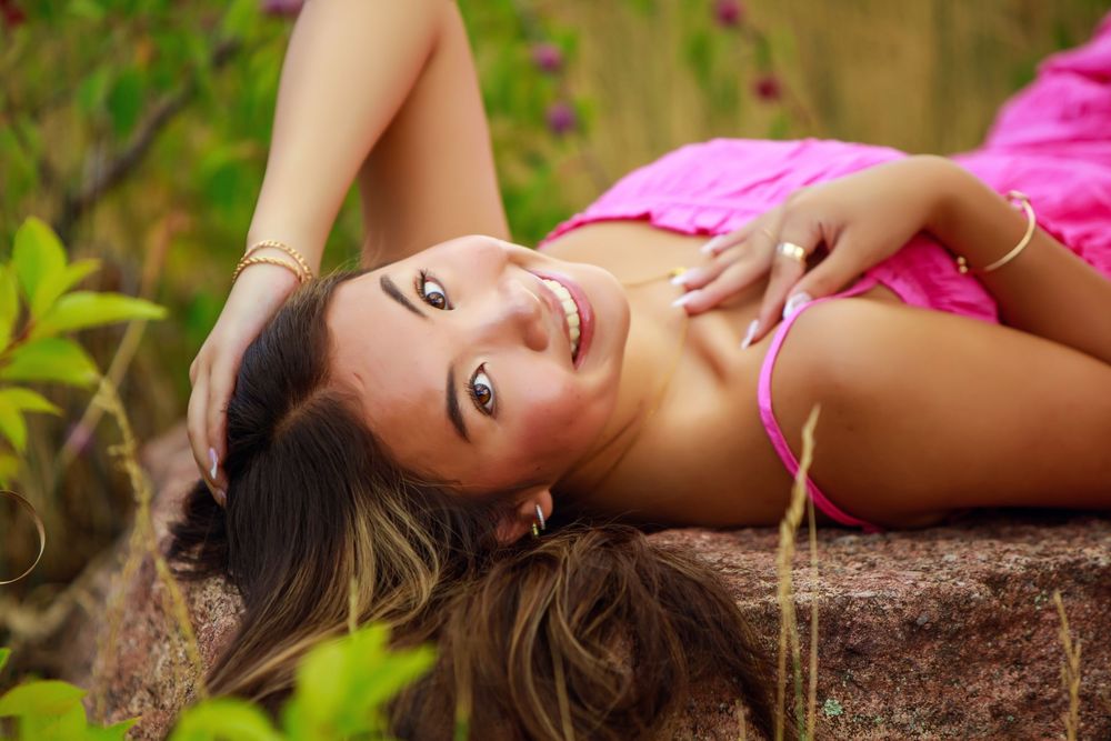 A person wearing a pink top laying in nature with greenery and flowers surrounding them in an outdoor portrait.