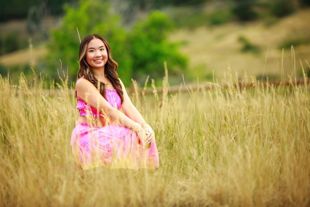 A cheerful woman in a pink flowy dress sits among tall golden grass in a summer field.
