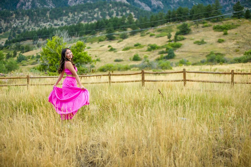 A vibrant pink dress twirls against golden grass with dramatic mountain peaks in the background.