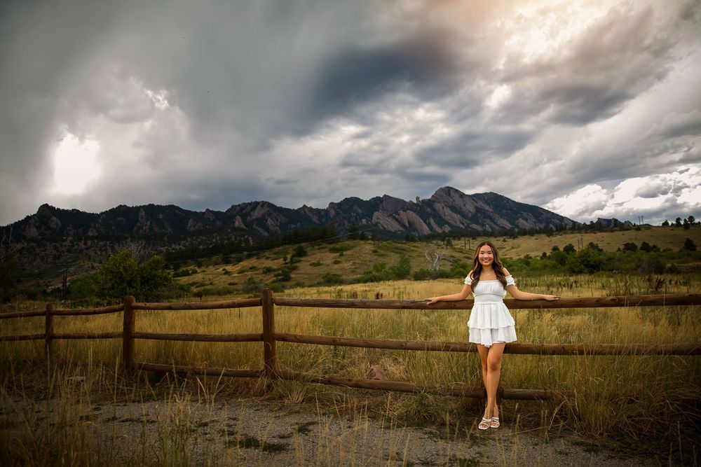 A woman in white dress stands by rustic fence with dramatic mountain landscape and stormy sky backdrop.