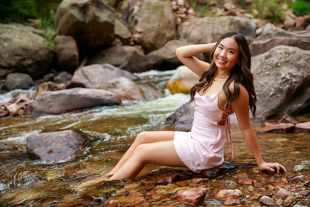 Someone in a white sundress sits on rocky terrain by a flowing stream on a sunny afternoon.