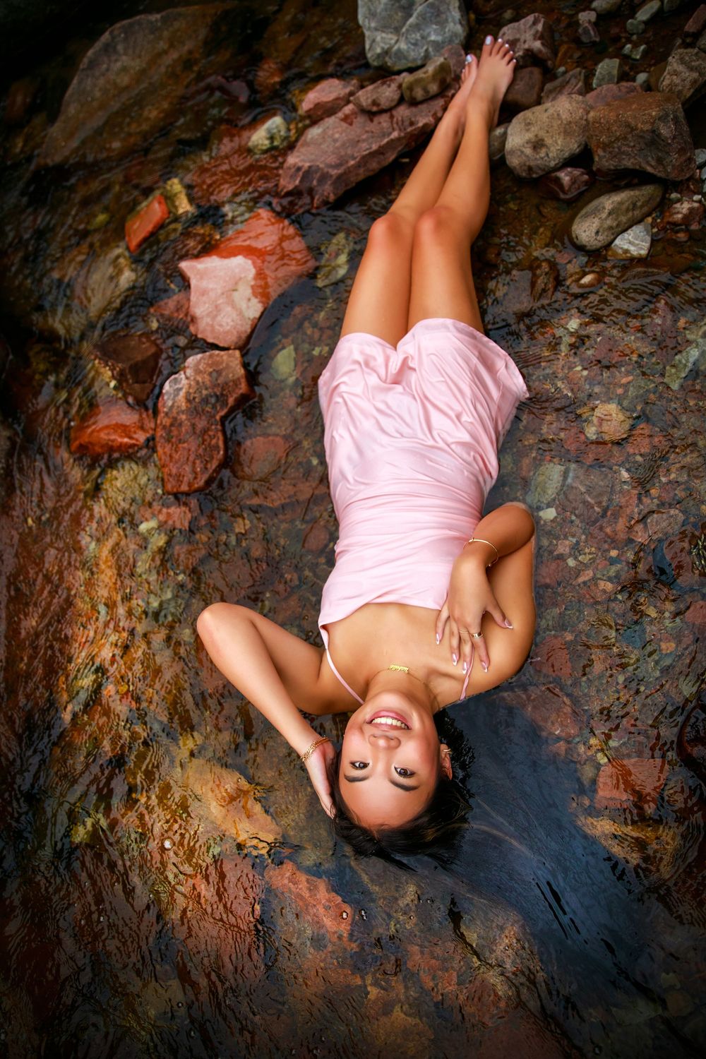 A model poses laying on textured rocks wearing a pink dress in an artistic fashion photography shot.